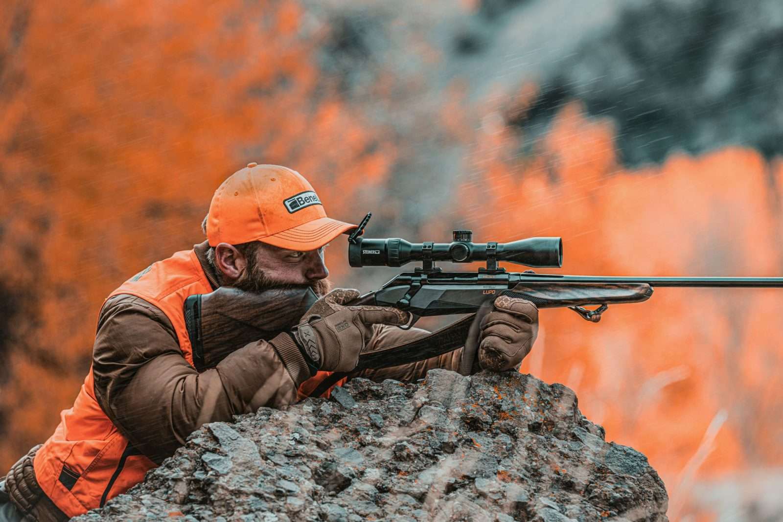 Hunter in blaze orange shouldering a scoped Benelli Lupo rifle off a rock with autumn aspens behind him