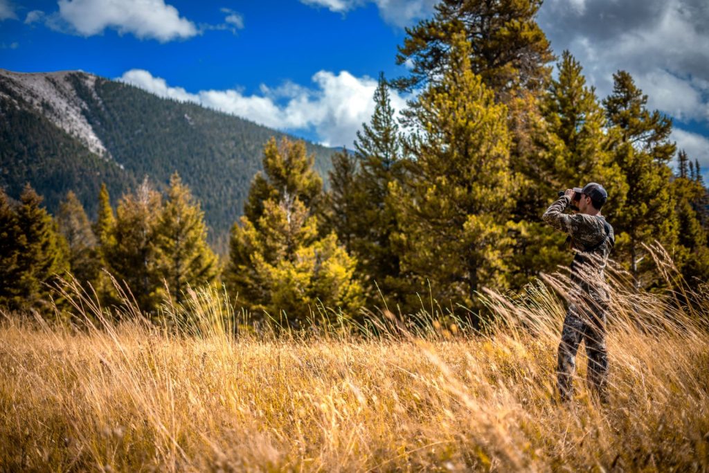 A hunter in camouflage glassing a mountain timber edge with binoculars
